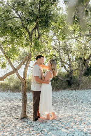 Couple embracing under green trees on sandy beach, woman in white dress and man in casual attire, golden sunlight filtering through.