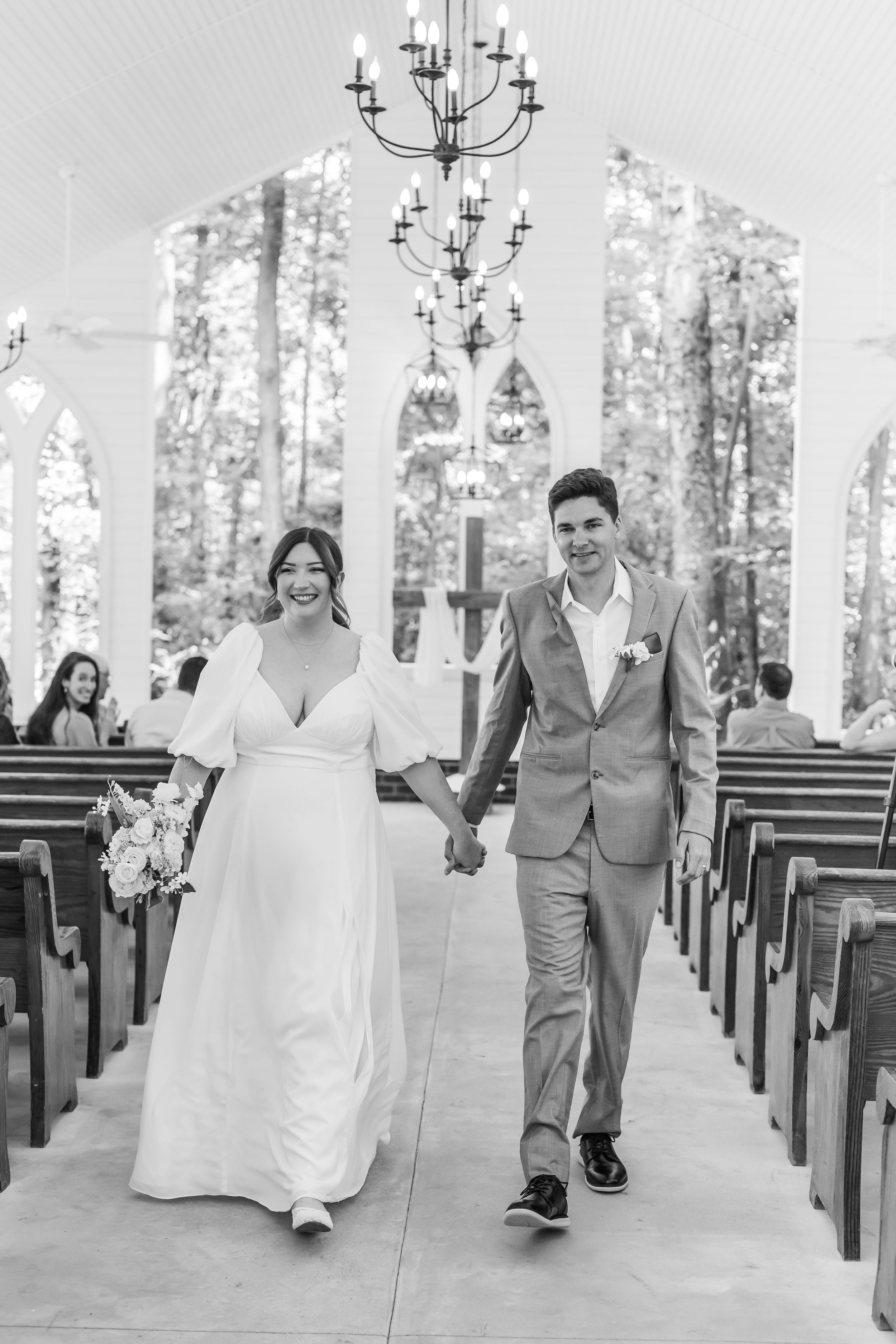 A joyful couple walks down the aisle of an outdoor chapel surrounded by trees.