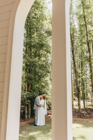 A romantic moment captured through white archway columns with lush forest backdrop.