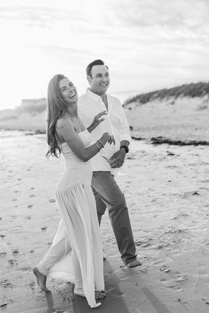 A joyful couple embraces while walking on a scenic beach in black and white.