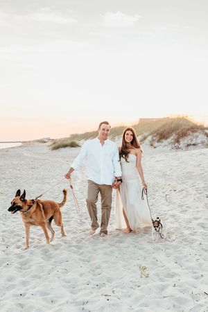 A couple walks their dogs along a sandy beach at sunset with coastal dunes in the background.