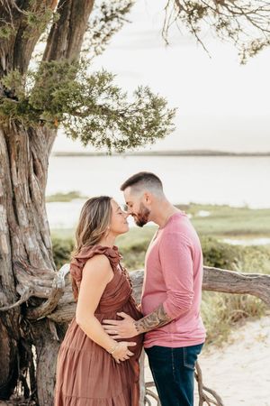 A couple shares tender moments during a maternity photoshoot on a beach with a large tree in the background.