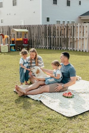 Family with two kids and a golden dog enjoying a backyard picnic on a blanket with watermelon on a sunny day.
