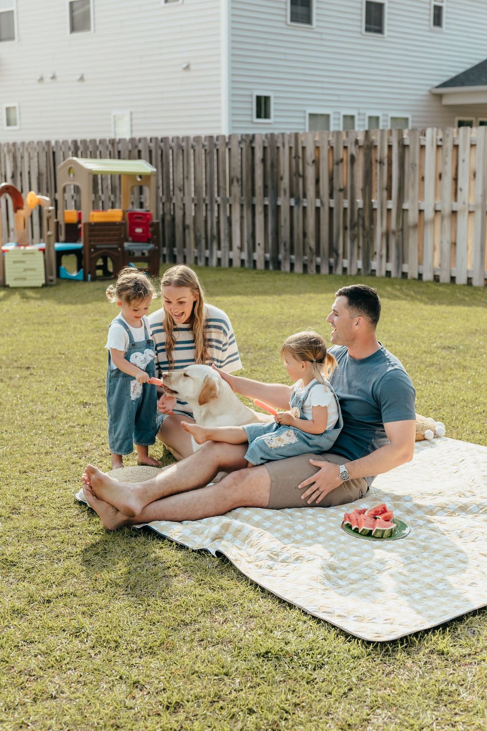 Family with two kids and a golden dog enjoying a backyard picnic on a blanket with watermelon on a sunny day.
