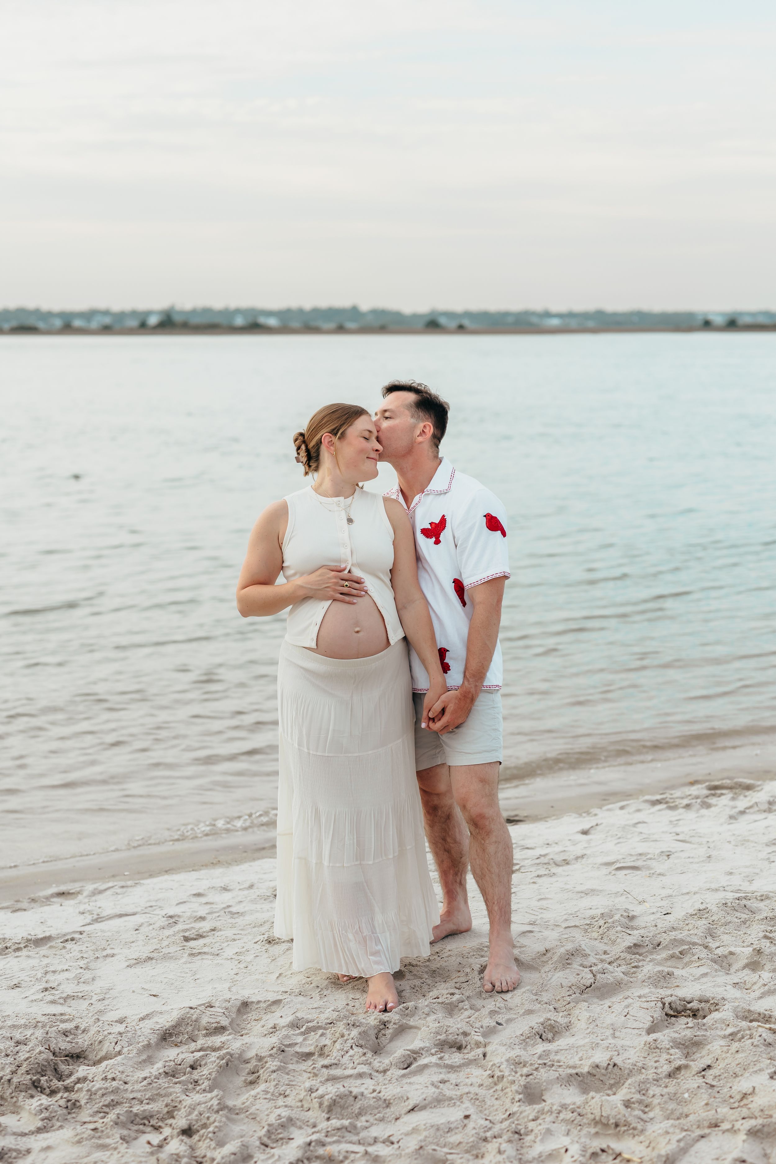 Pregnant couple sharing a tender kiss on a sandy beach, wearing white outfits with calm ocean water in the background.