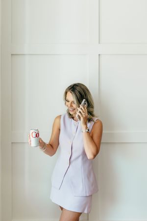 Woman in lavender dress smiling while talking on phone and holding a beverage can against white wall.