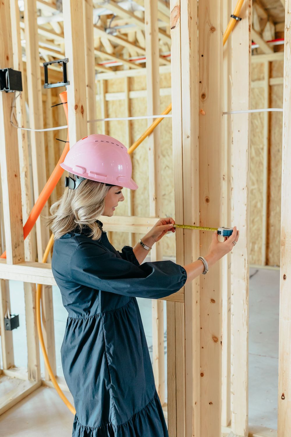 Woman in pink hard hat measuring wooden wall framing at a residential construction site.