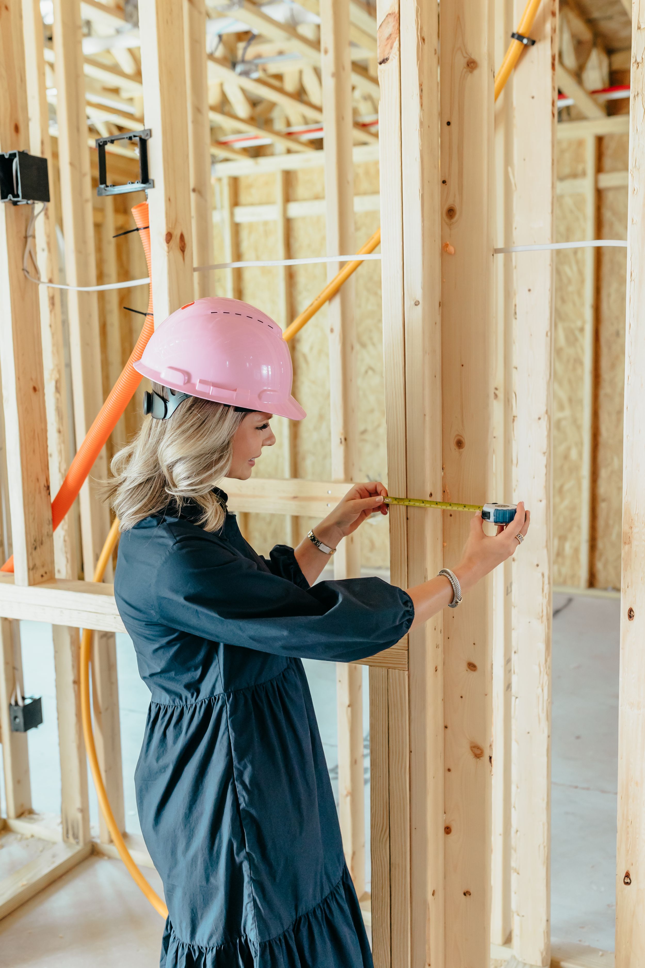 Woman in pink hard hat measuring wooden wall framing at a residential construction site.