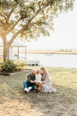 Family enjoying a sunset moment together by the water under a large tree with a dock in the background.