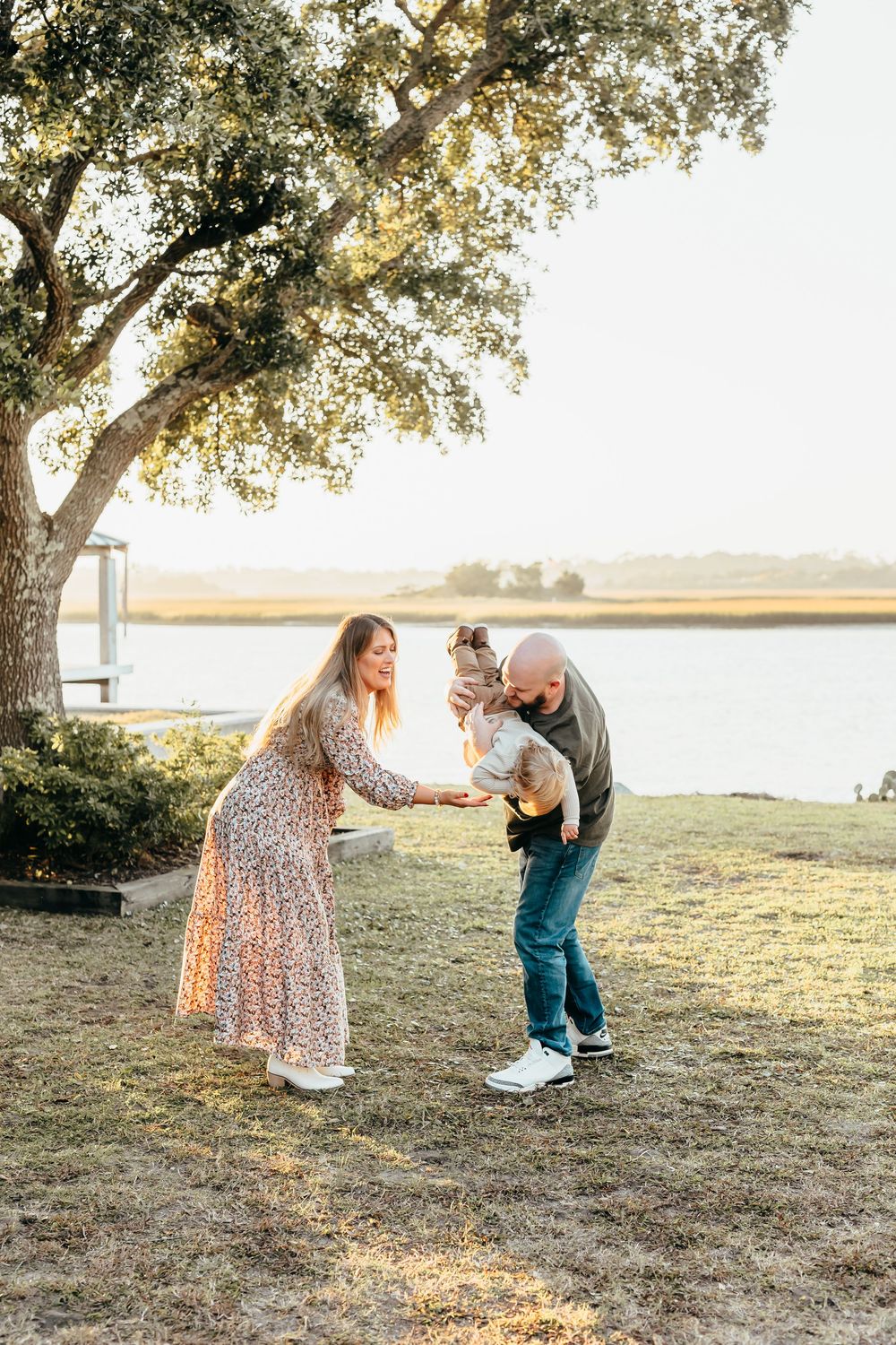 A couple playing with a small child under a large tree by a serene waterfront during golden hour.