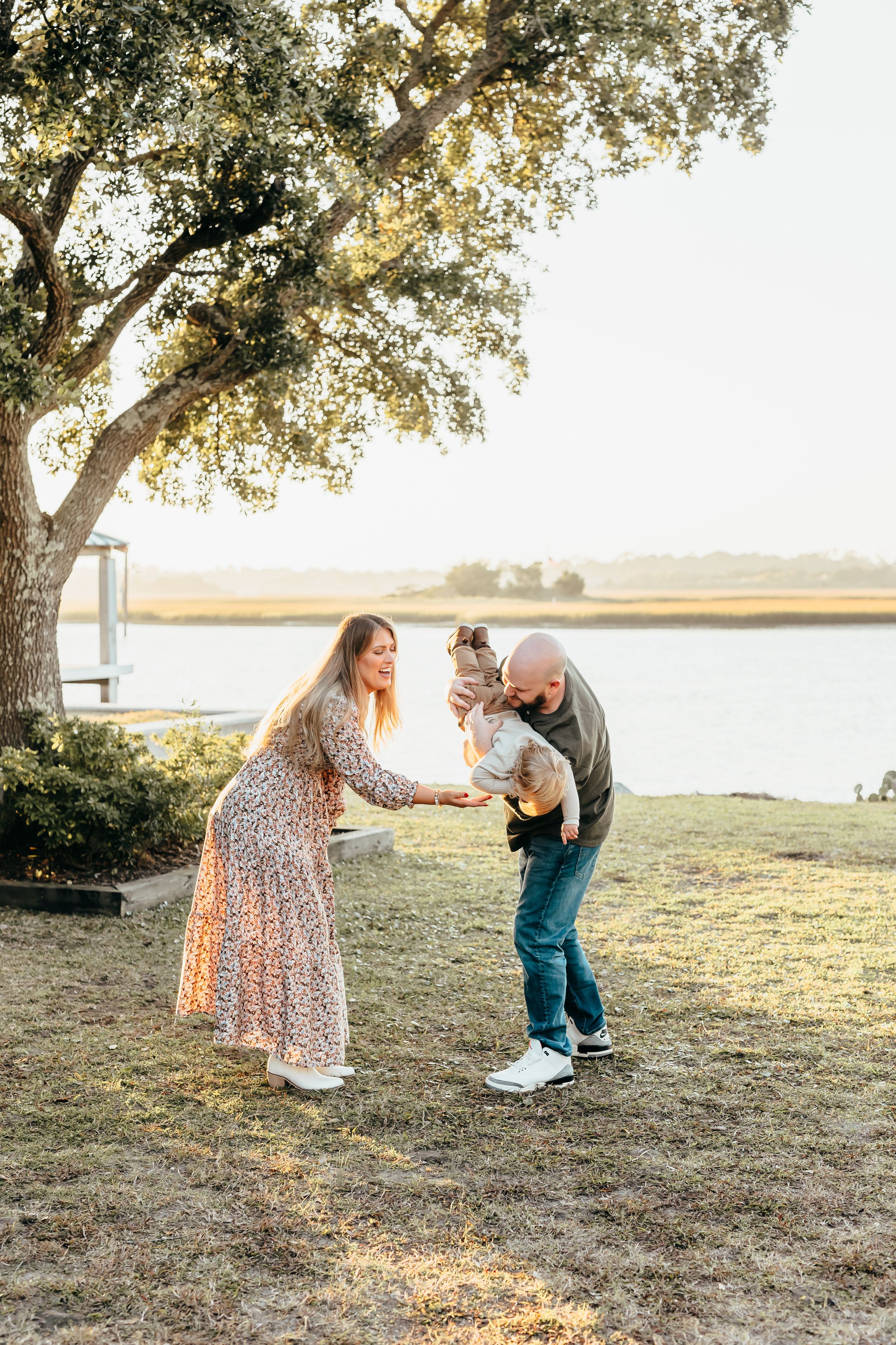 A couple playing with a small child under a large tree by a serene waterfront during golden hour.