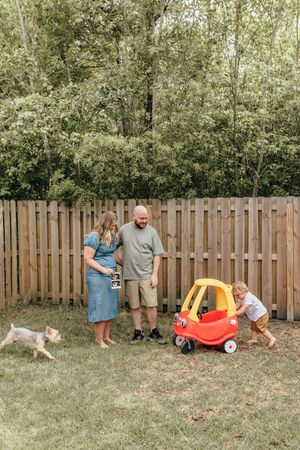 Family with toddler and dog playing in backyard with Little Tikes car, holding ultrasound photo for pregnancy announcement.