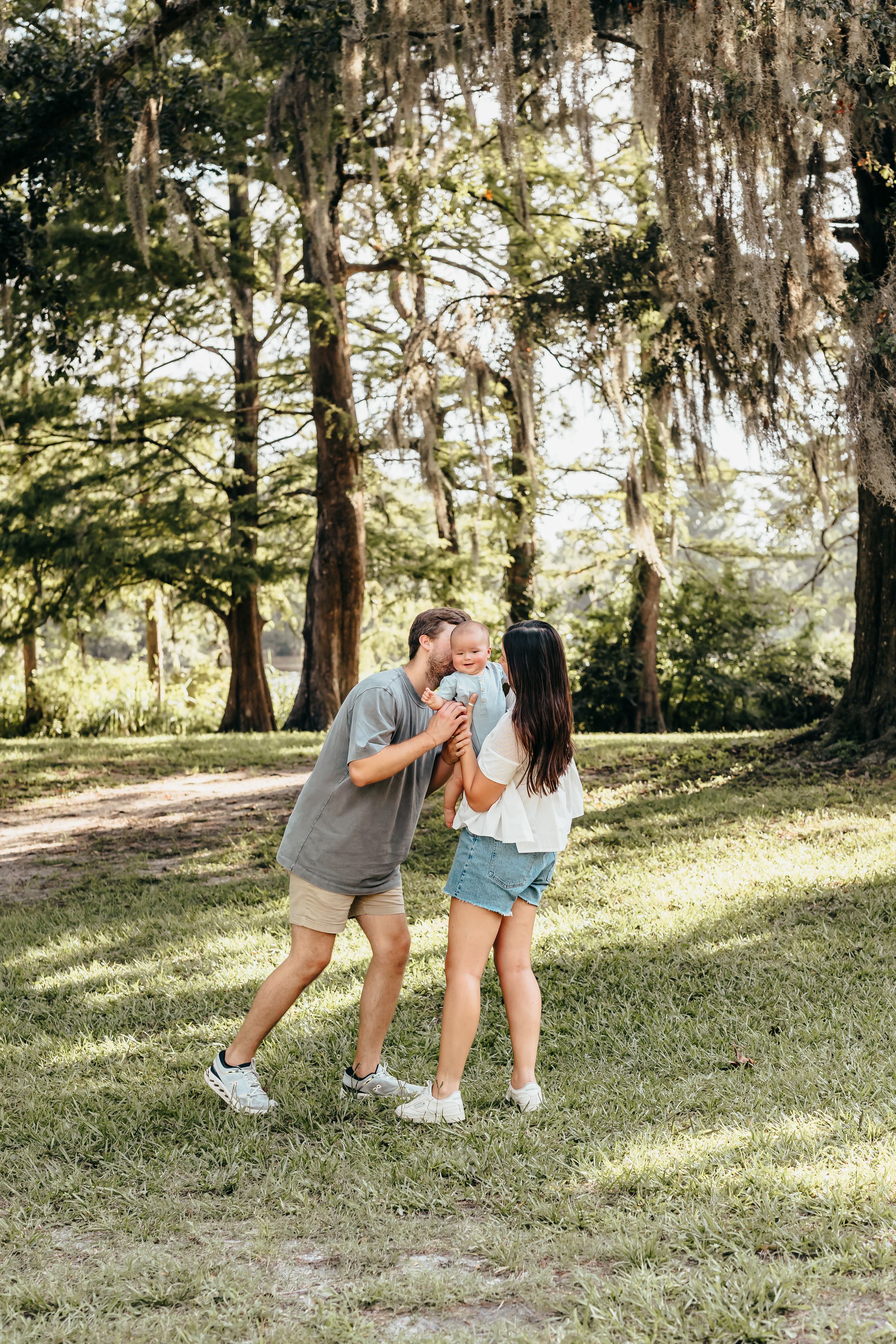 A family playfully dancing together in a sun-dappled park with Spanish moss hanging from the trees in the background. Wilmington NC