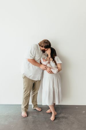 A couple in white and beige clothing share a kiss while cradling their newborn baby against a white wall.
