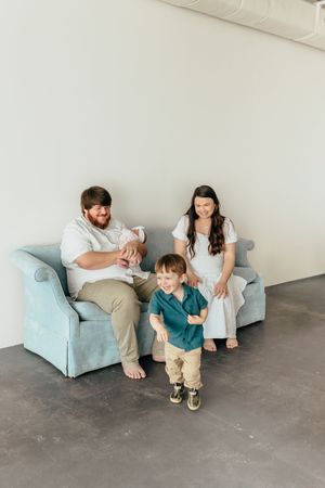 A young family with a newborn and toddler poses on a light blue sofa in a bright studio setting.