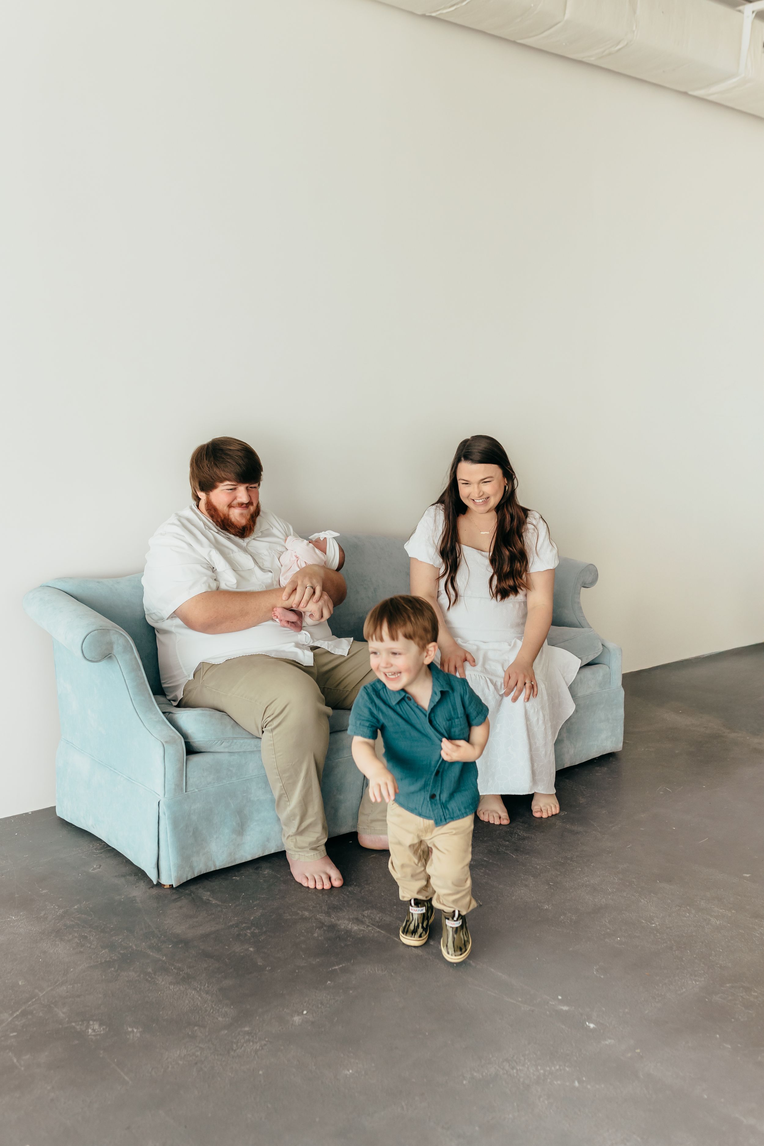 A young family with a newborn and toddler poses on a light blue sofa in a bright studio setting.