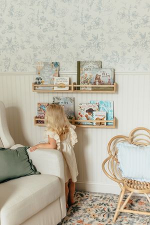 Young girl in white dress browses colorful children's books on gold wall-mounted shelves in a cozy nursery room.