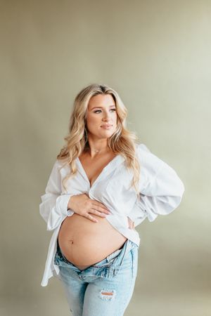 An expectant woman in a white button-down shirt and distressed jeans poses against a neutral backdrop.