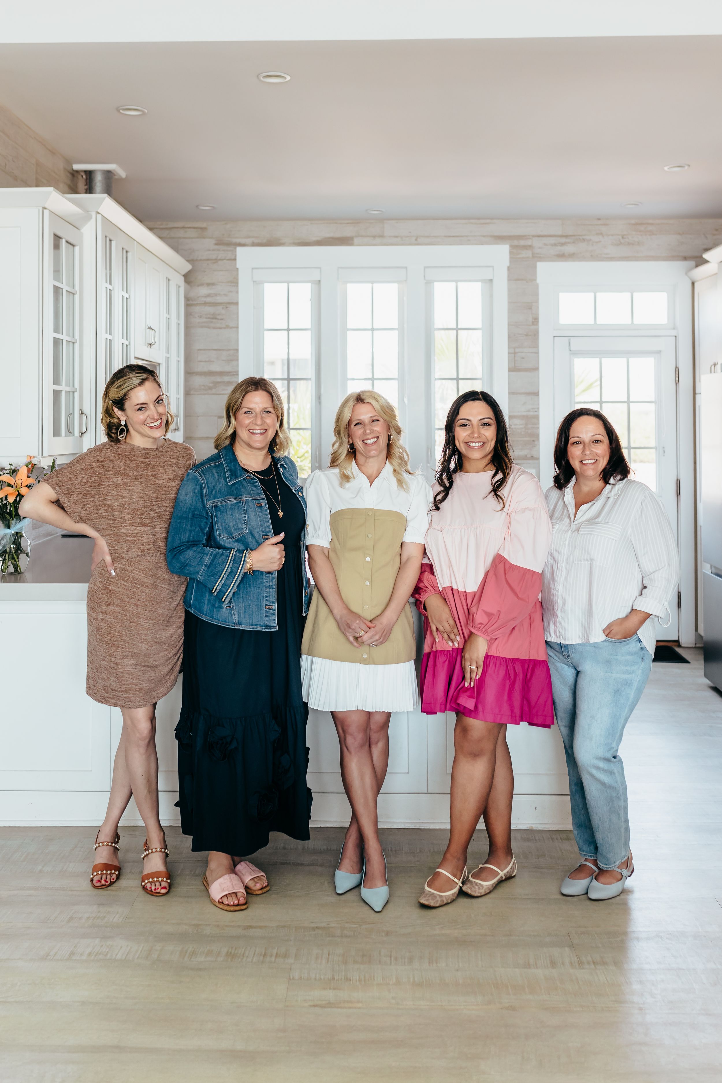 Five women smiling together indoors, wearing colorful spring outfits including dresses and casual tops.