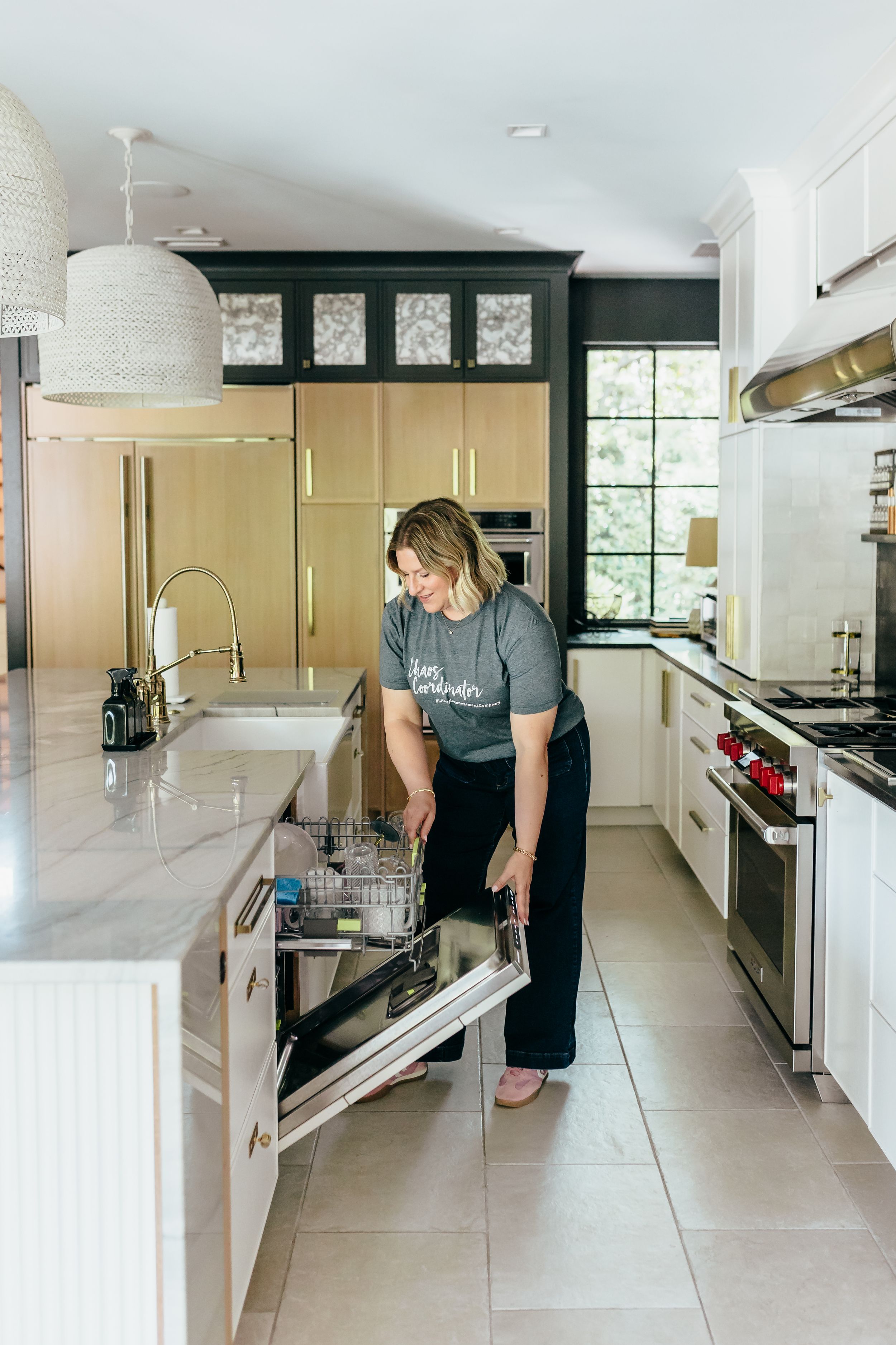 Woman loading dishwasher in modern kitchen with white cabinets, black accents, marble countertops, and stainless steel appliances.