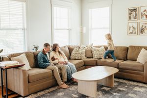 Family relaxing together on brown leather couch in bright living room with natural light streaming through windows.