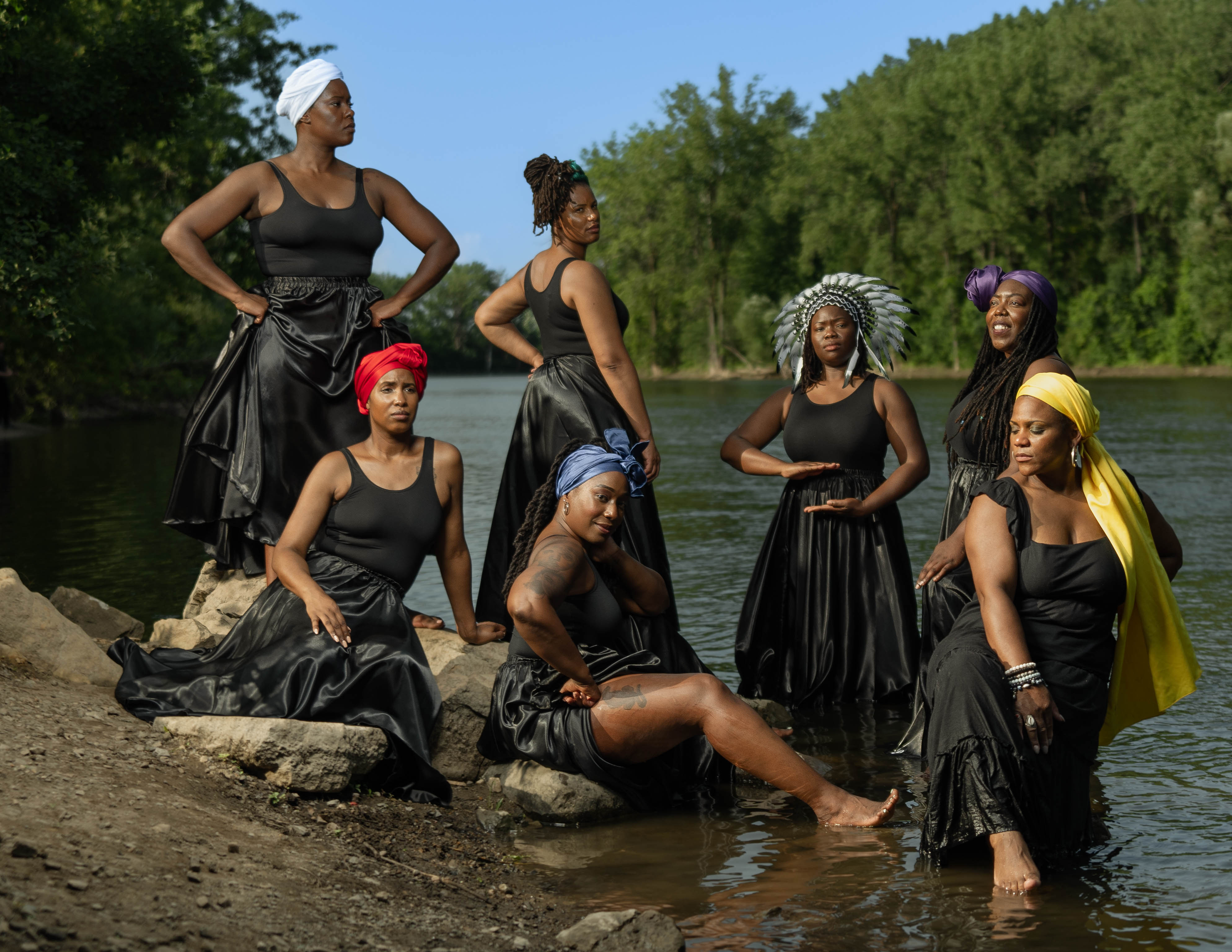 Group of seven Black women in black dresses posing by a river in a nature setting.