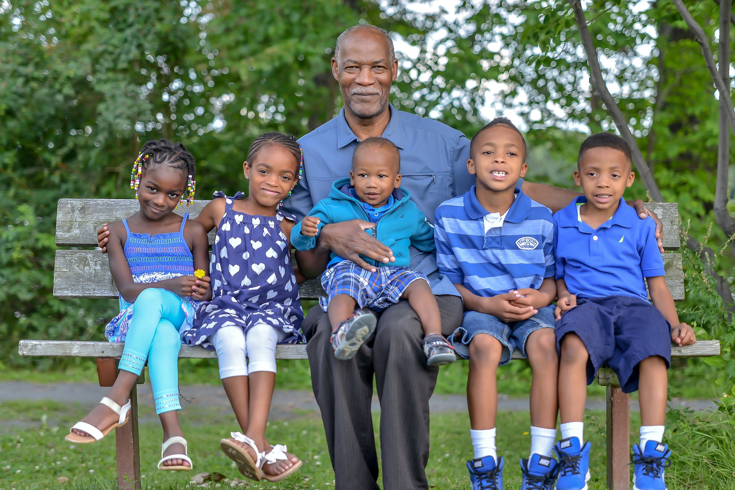 Elderly Black grandfather sitting on park bench with five young grandchildren dressed in blue outfits.