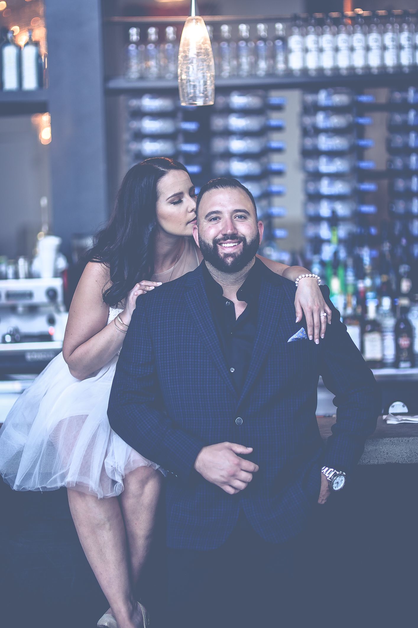 A woman in a white dress kisses a smiling bearded man on the cheek at an elegant bar setting.