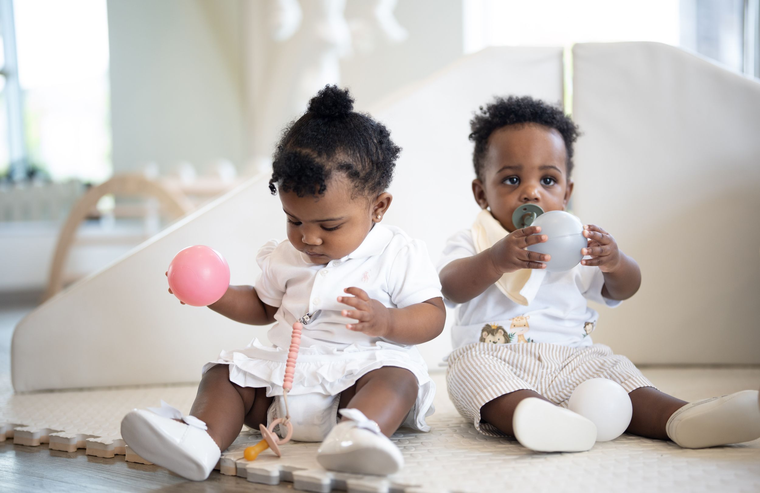 Two adorable baby toddlers sit on floor playing with toys in bright modern nursery room.