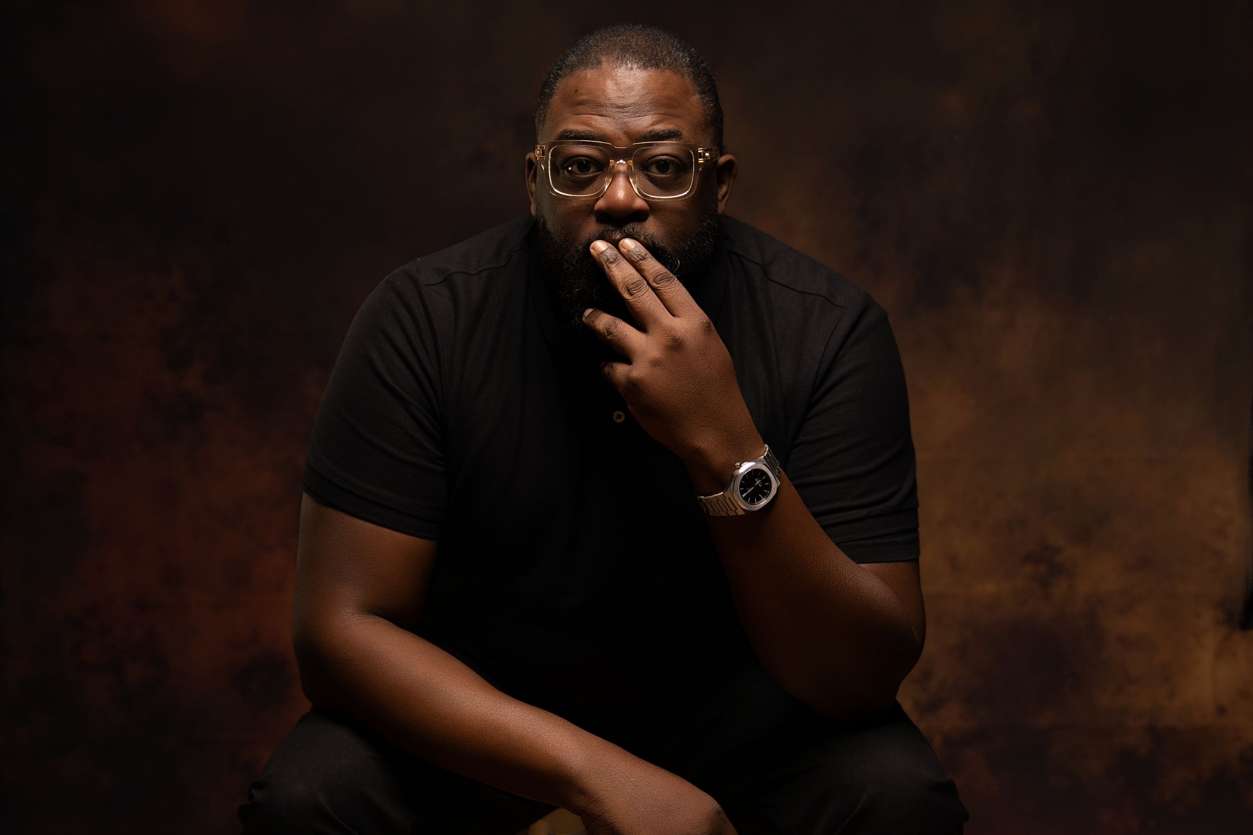 Man in black outfit with glasses sitting thoughtfully against textured brown background.