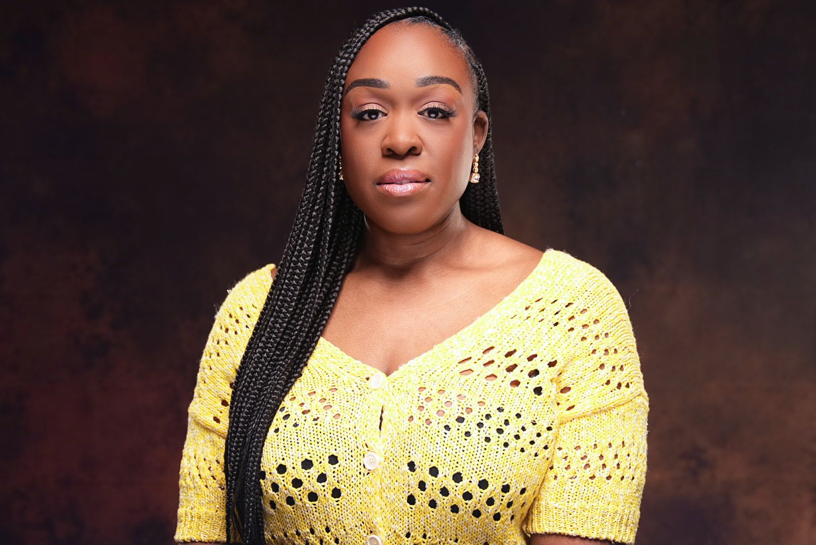 Woman with long braids in yellow crochet top posing against warm brown background.