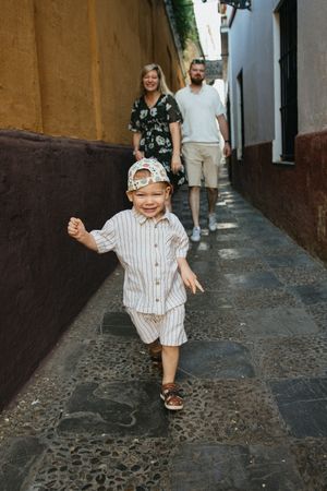 A toddler in striped outfit runs joyfully down a narrow cobblestone alley between old stone walls.