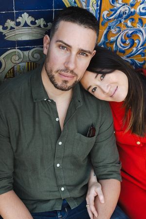 A couple in olive green and red outfits pose together against a decorative blue and gold tiled wall.