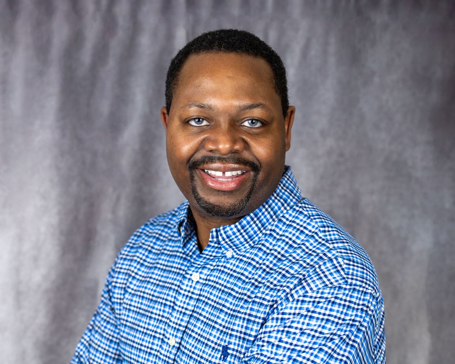 Professional headshot of a person in a blue checkered shirt smiling against gray background.