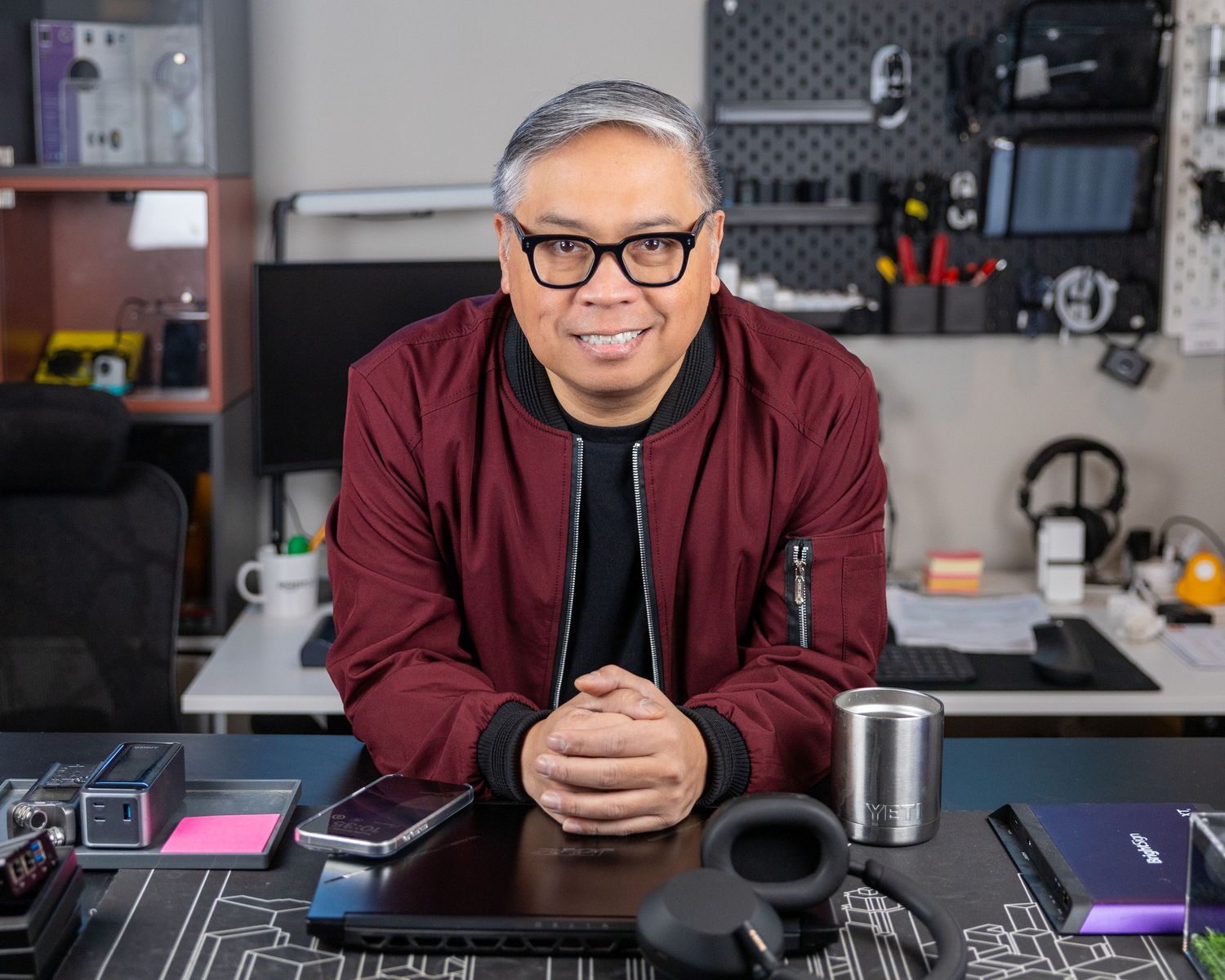 Person in glasses and burgundy jacket sitting at desk with tech equipment and coffee.