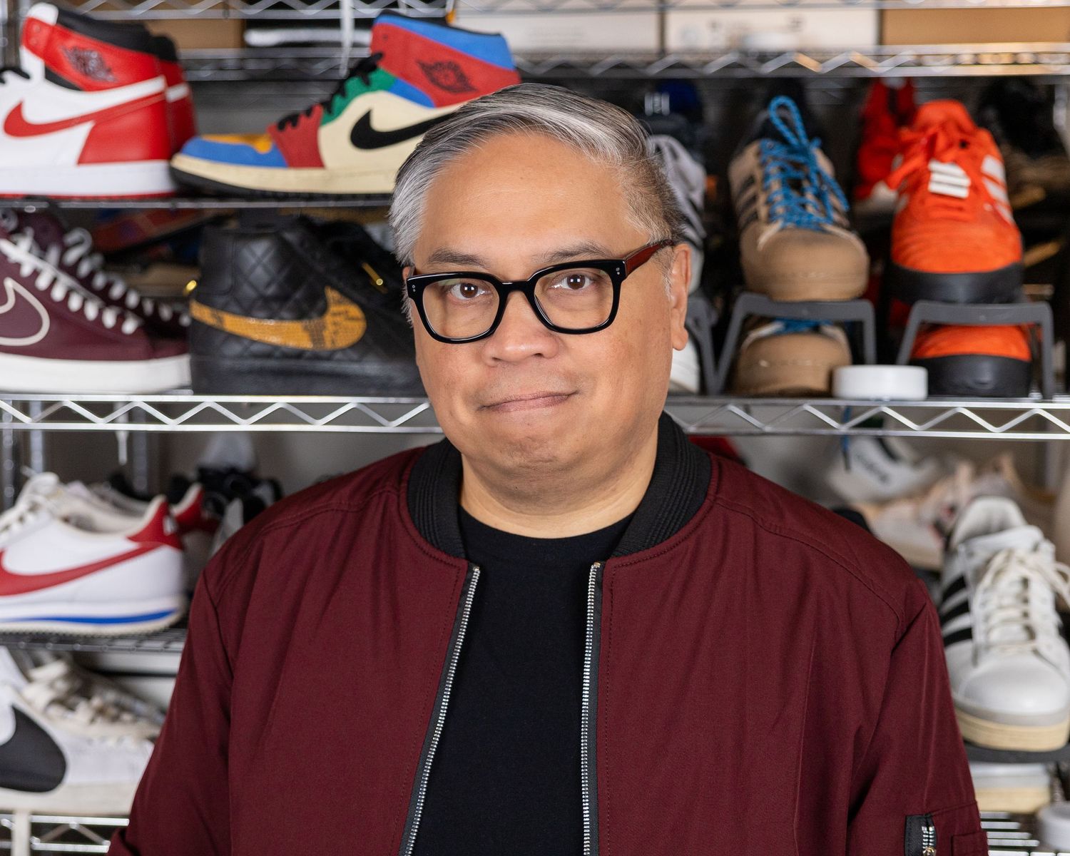 Person in burgundy jacket and glasses photographed in front of shelves displaying sneaker collection.