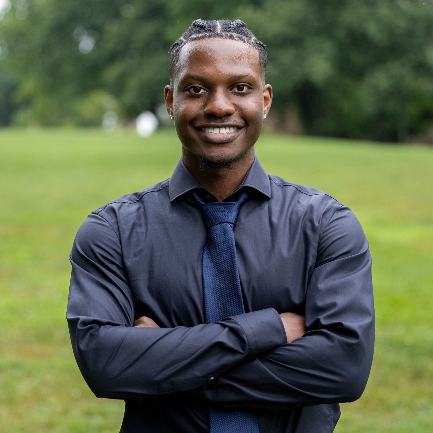 Professional portrait of someone in a navy suit and tie smiling warmly against a green outdoor background.