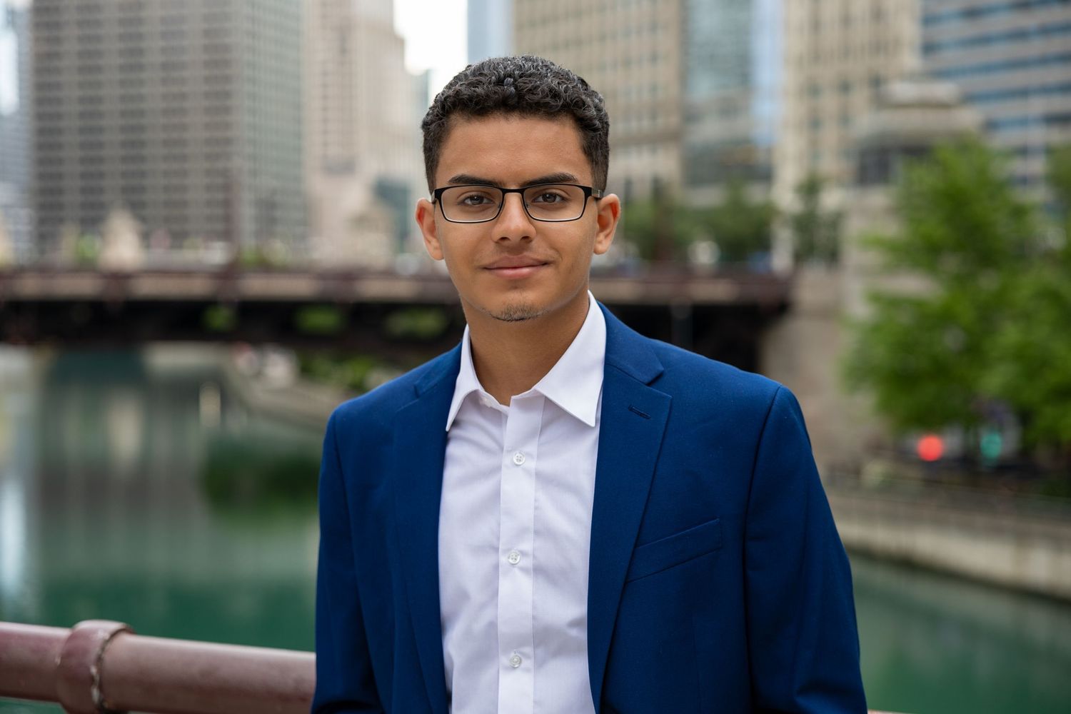 Professional headshot of a person in a blue suit and glasses against an urban waterfront background.