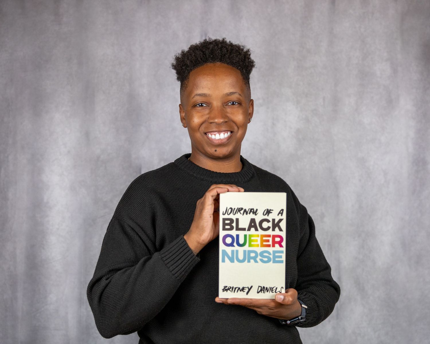Person in black sweater holding book titled 'Black Queer Nurse' with joyful expression.