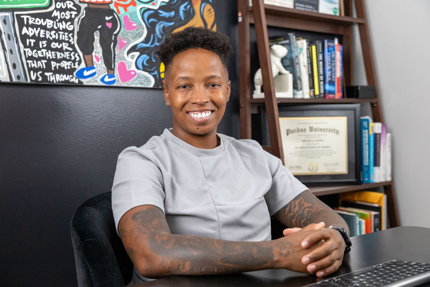 Person sitting at desk with laptop and bookshelf in background displaying warm smile.