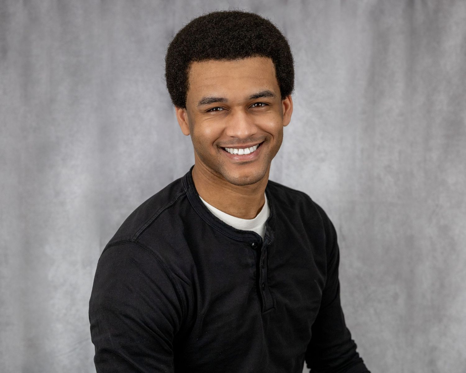 Professional portrait of person in black jacket with bright smile against gray background.