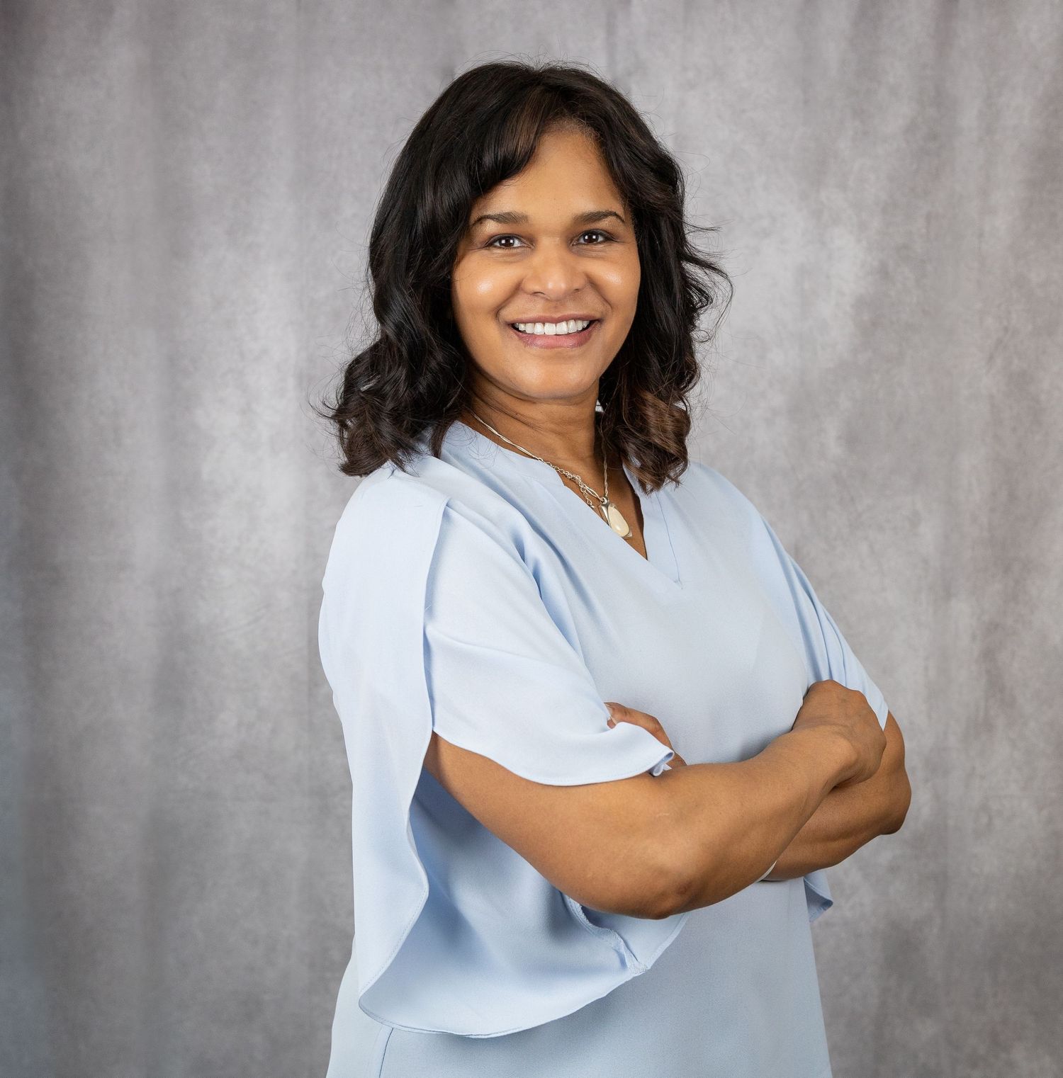 Healthcare worker in light blue scrubs poses with crossed arms against gray backdrop.