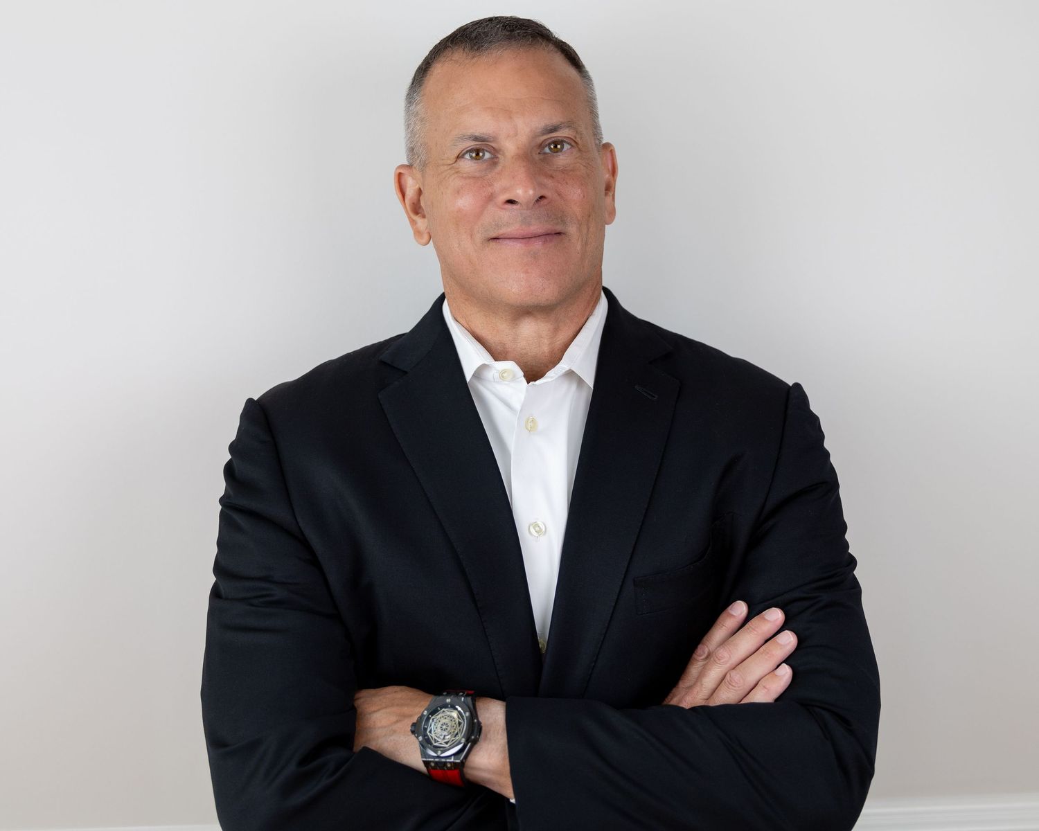 Executive in dark suit and white shirt poses with crossed arms against white background.