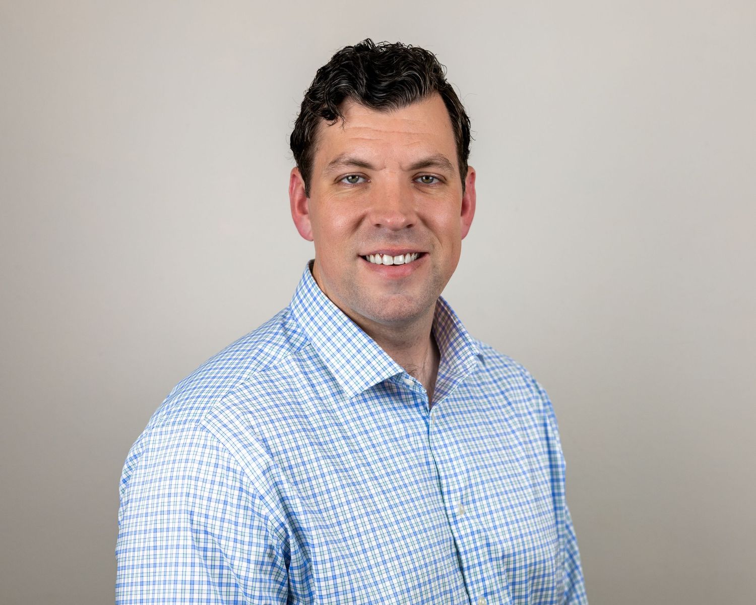Professional headshot of person in blue checkered dress shirt against neutral background.