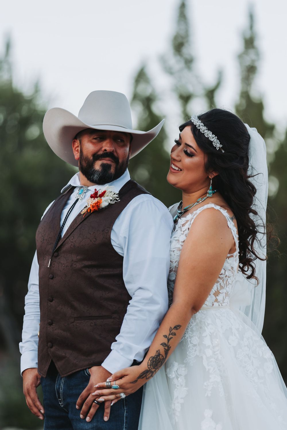A western-themed wedding couple poses outdoors with the bride in white and groom in a brown vest and cowboy hat.