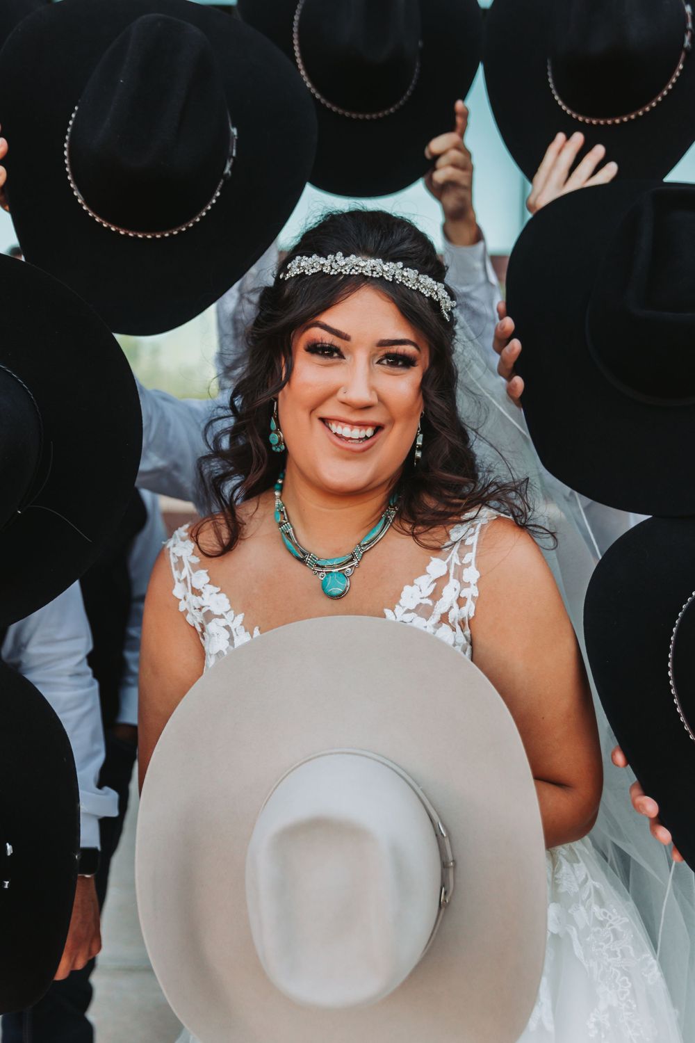 A bride in lace wearing a crystal headpiece smiles surrounded by cowboy hats.