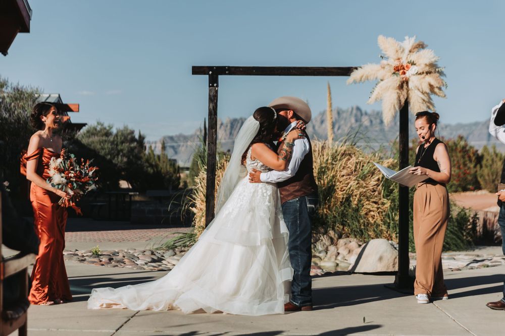 A wedding ceremony takes place under a wooden arch against a desert mountain backdrop.