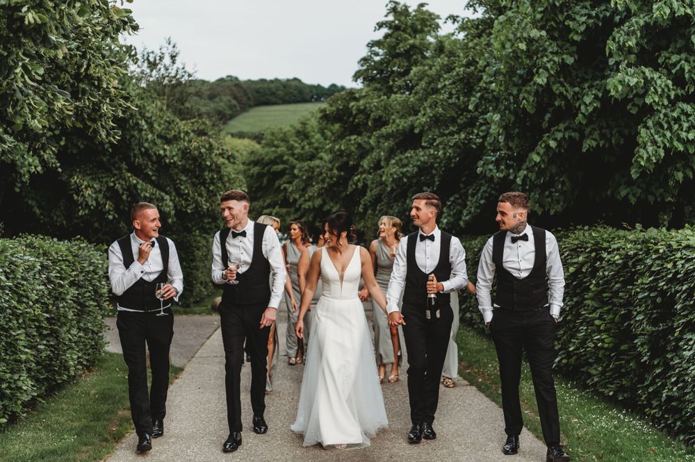 Wedding party walking along garden path with tall hedges on sunny day.