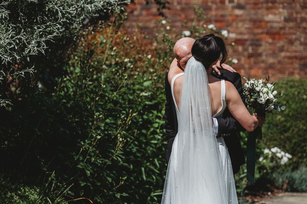 Couple shares intimate moment in garden setting with brick wall and white flowers.