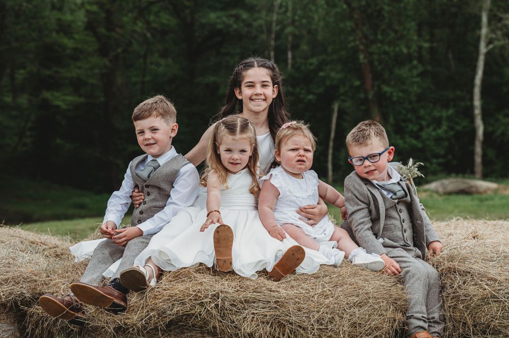 Children in formal attire sit together on a hay bale in front of a wooded background.